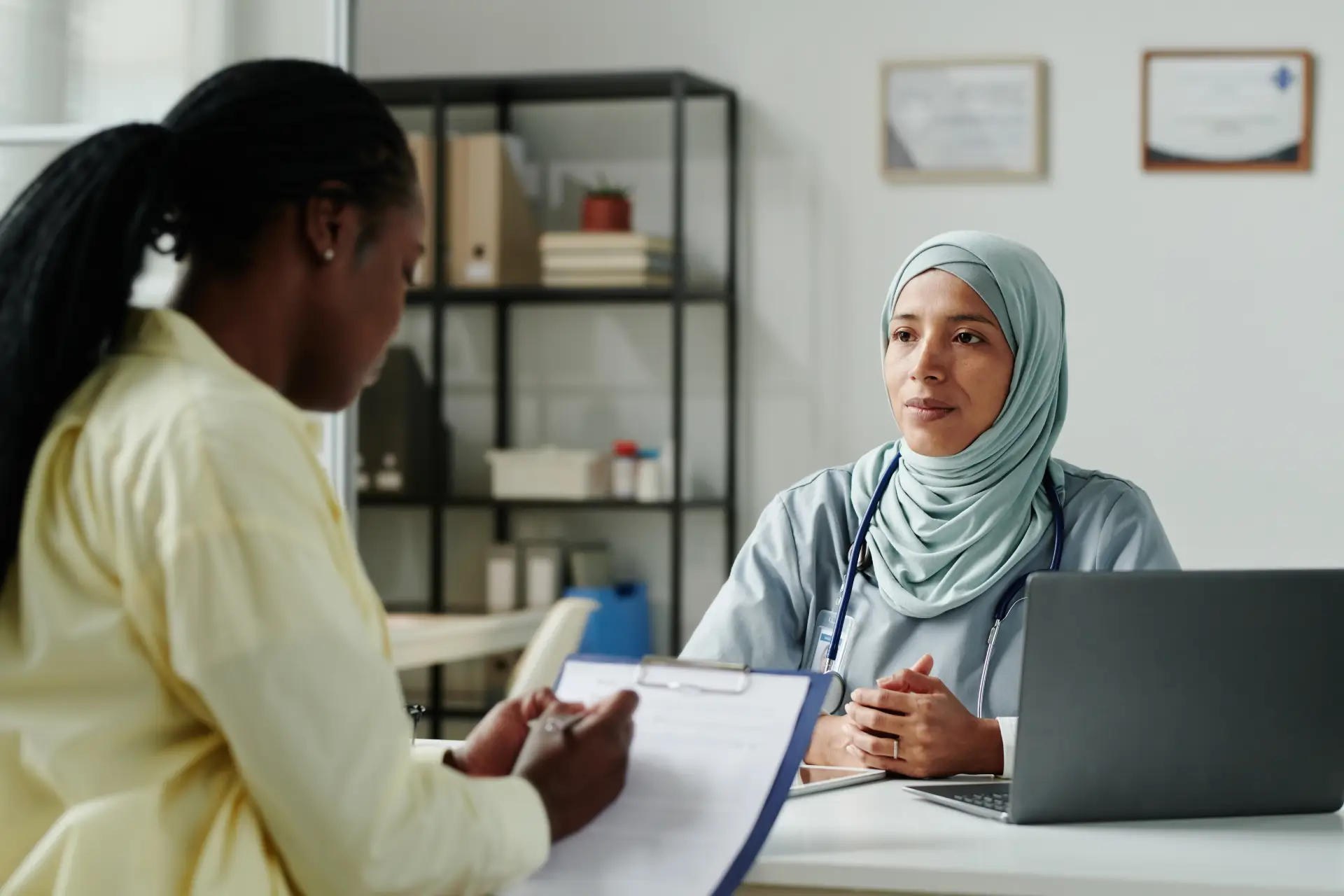 A woman fills out forms on a clipboard across a desk from a medical professional with a laptop on the desk and credentials hanging on the wall behind her