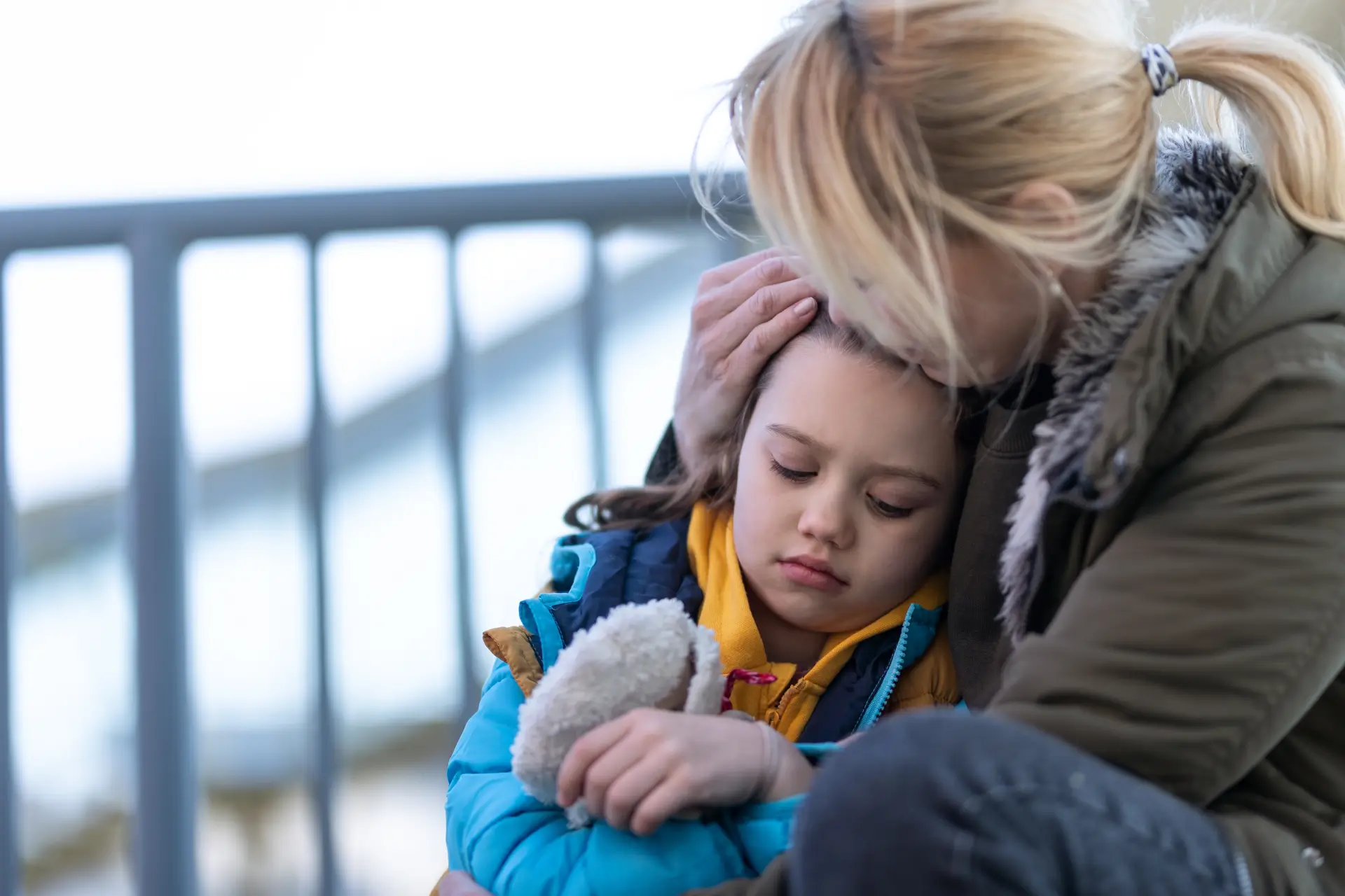 A parent hugs her child. They both look sad and are outside wearing coats.
