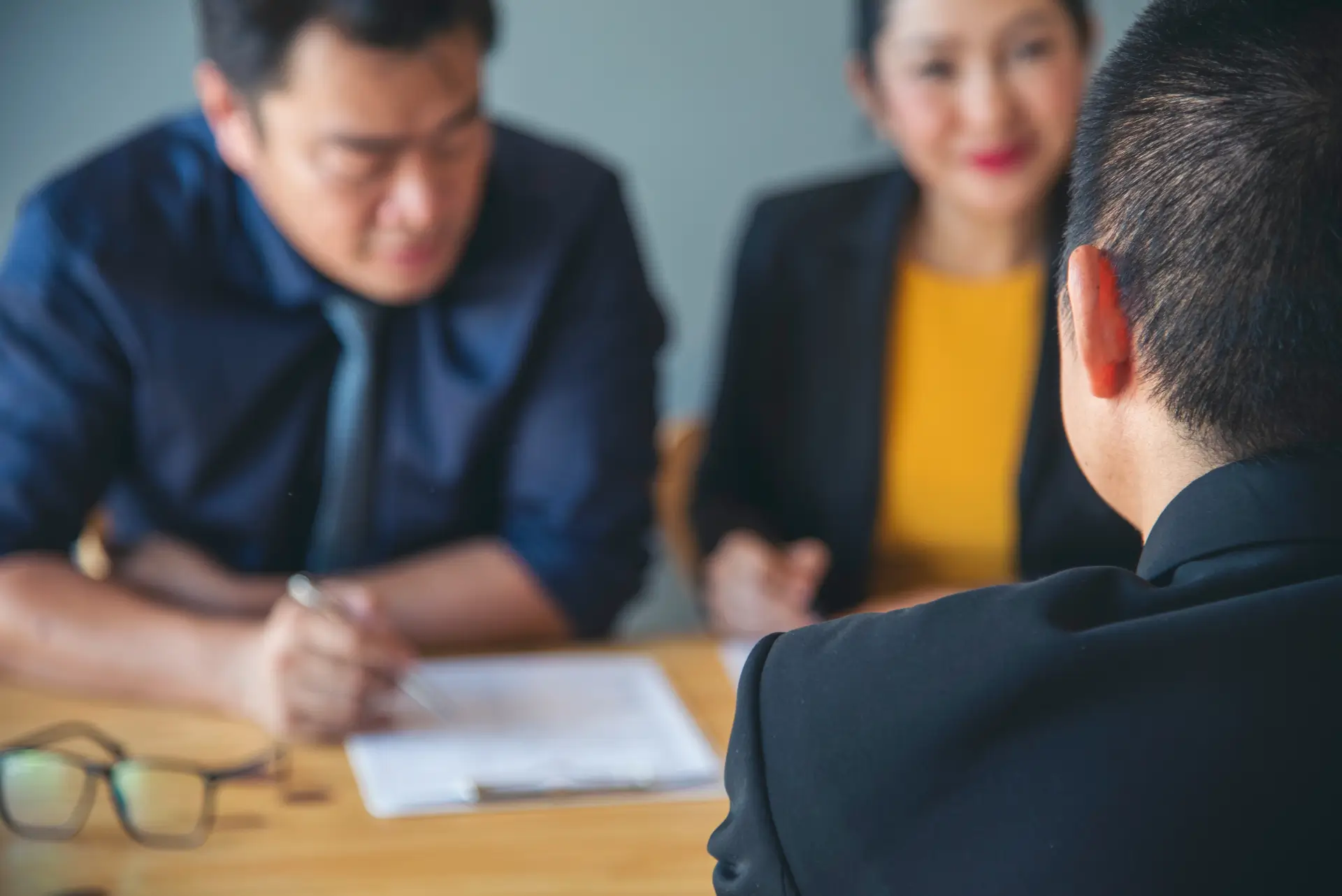 A man and woman sit across from a man, all dressed in business attire, speak to each other across a desk