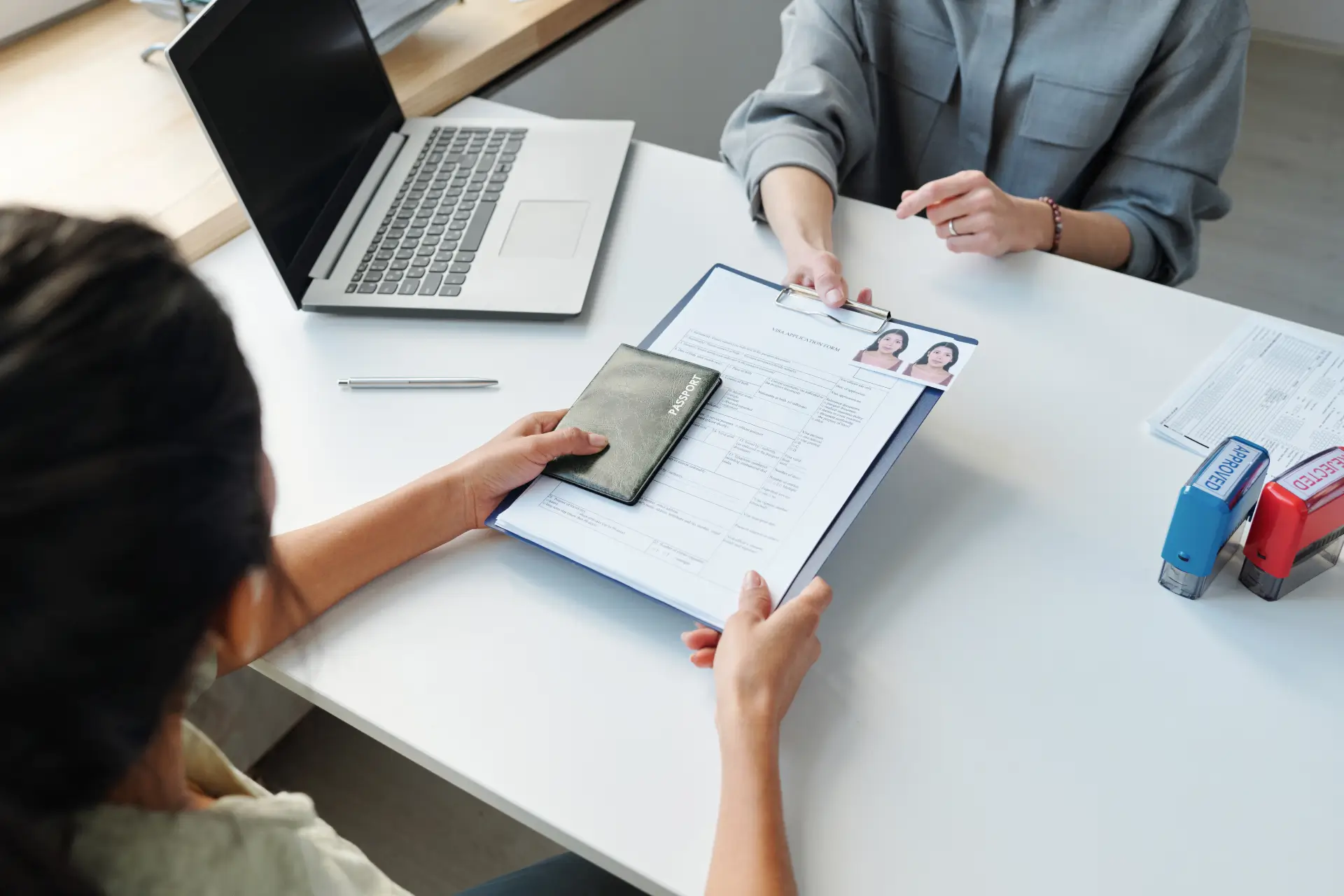 A woman hands another woman a clipboard with a passport application with passport pictures to another woman. On the desk there is a laptop and 2 stamps labeled: APPROVED and REJECTED