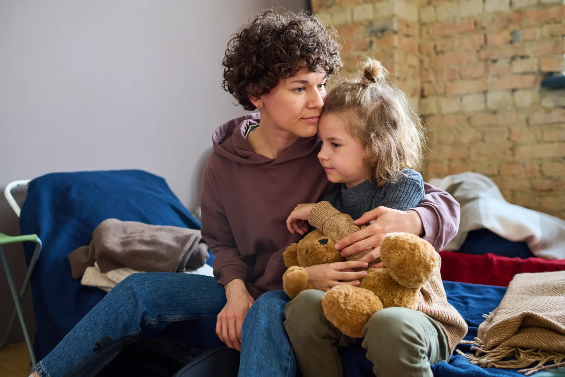 A woman hugs a little girl who is hugging a teddy bear, both sitting on a bed